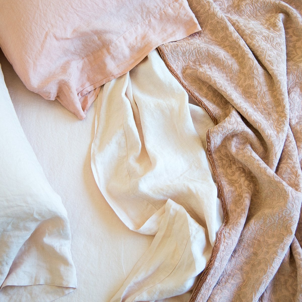 : Close-up of rumpled Vienna coverlet with linen sheets and sleeping pillows - in pink and cream tones - overhead view.