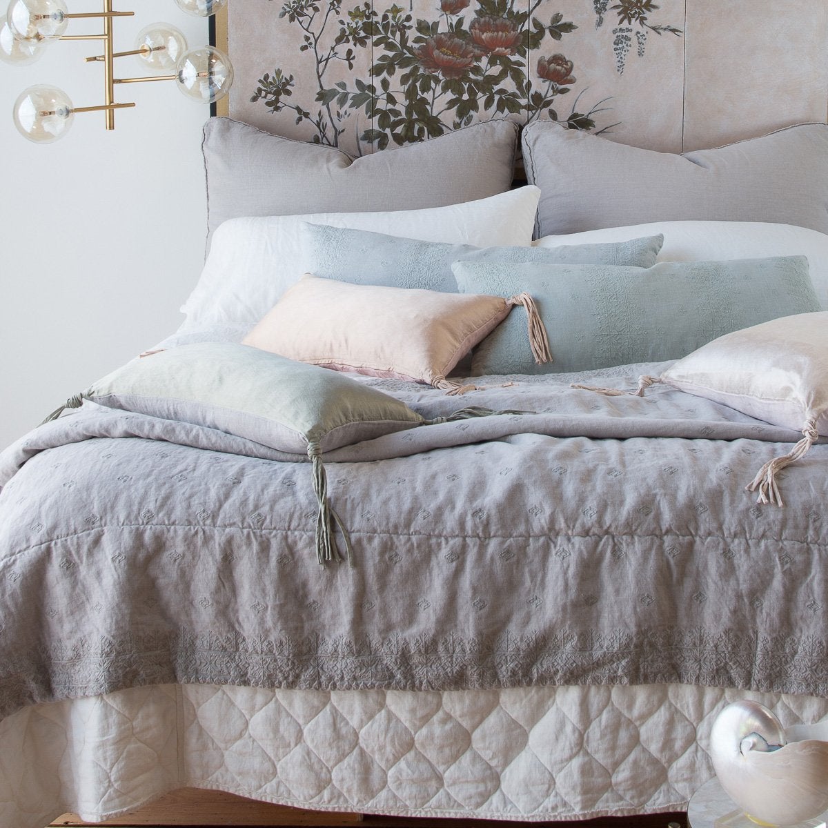 : Ines throw blanket layered with mixed textures of charmeuse, quilted linen, and plain linen in soft grey, blue, pink and white tones - end of bed view. 