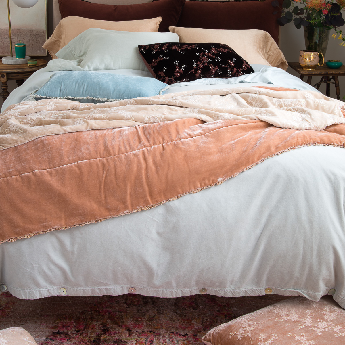 [allvariants]: Carmen throw blanket in rouge layered under silk velvet throw pillows and blankets in light neutrals, soft blues, and deep browns - end of bed view.