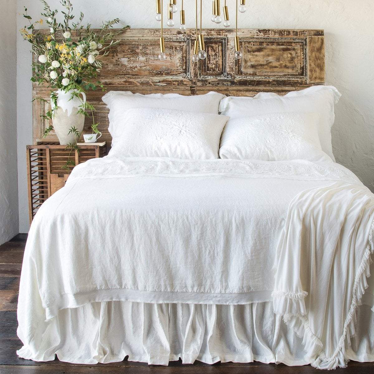[allvariants]: Delphine coverlet in white on an all-white beed with shams, sleeping pillows, throw blanket and bed skirt - end of bed view. 