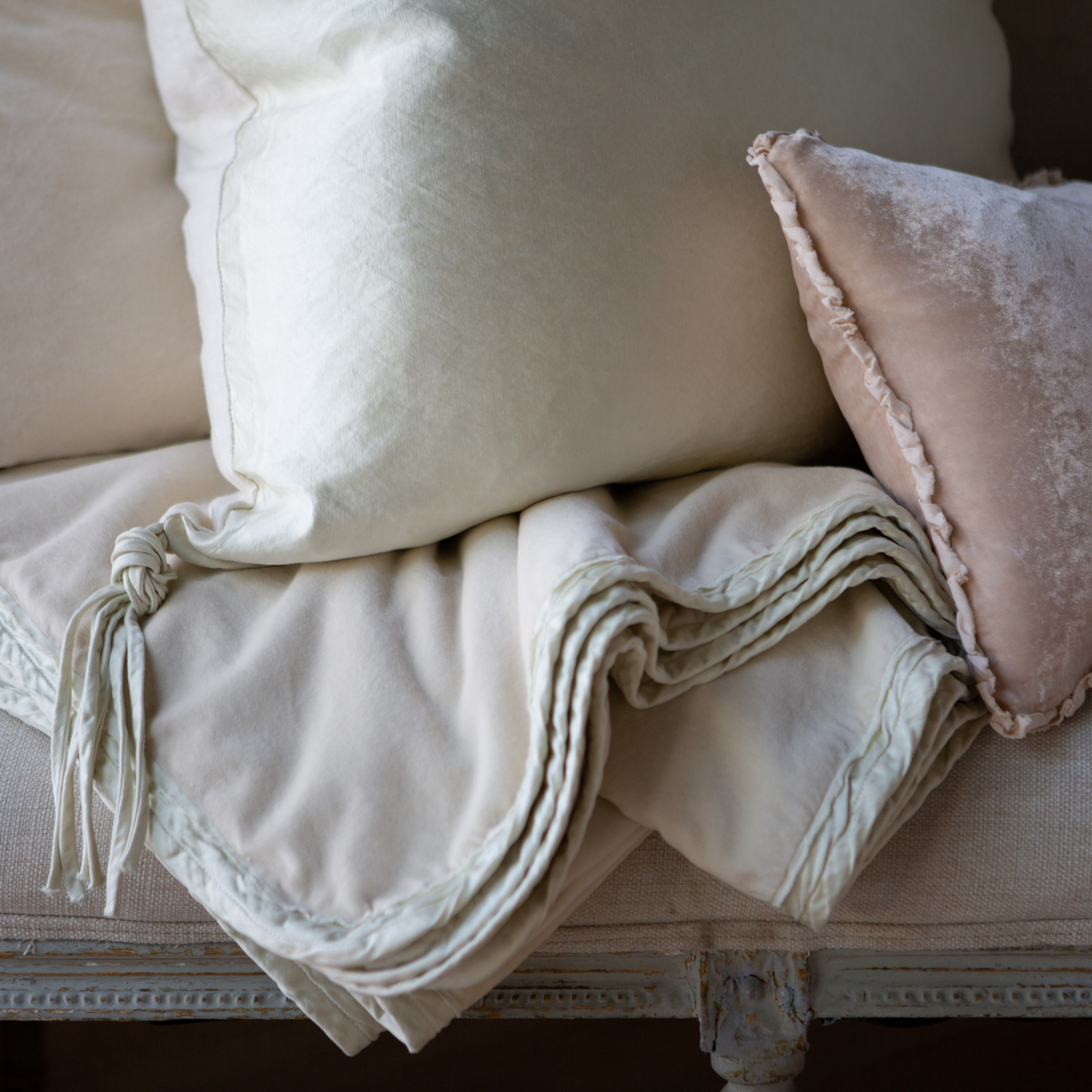 : Close-up of Harlow throw blanket in parchment folded under light neutral throw pillows, showcasing shining carmeuse trim.