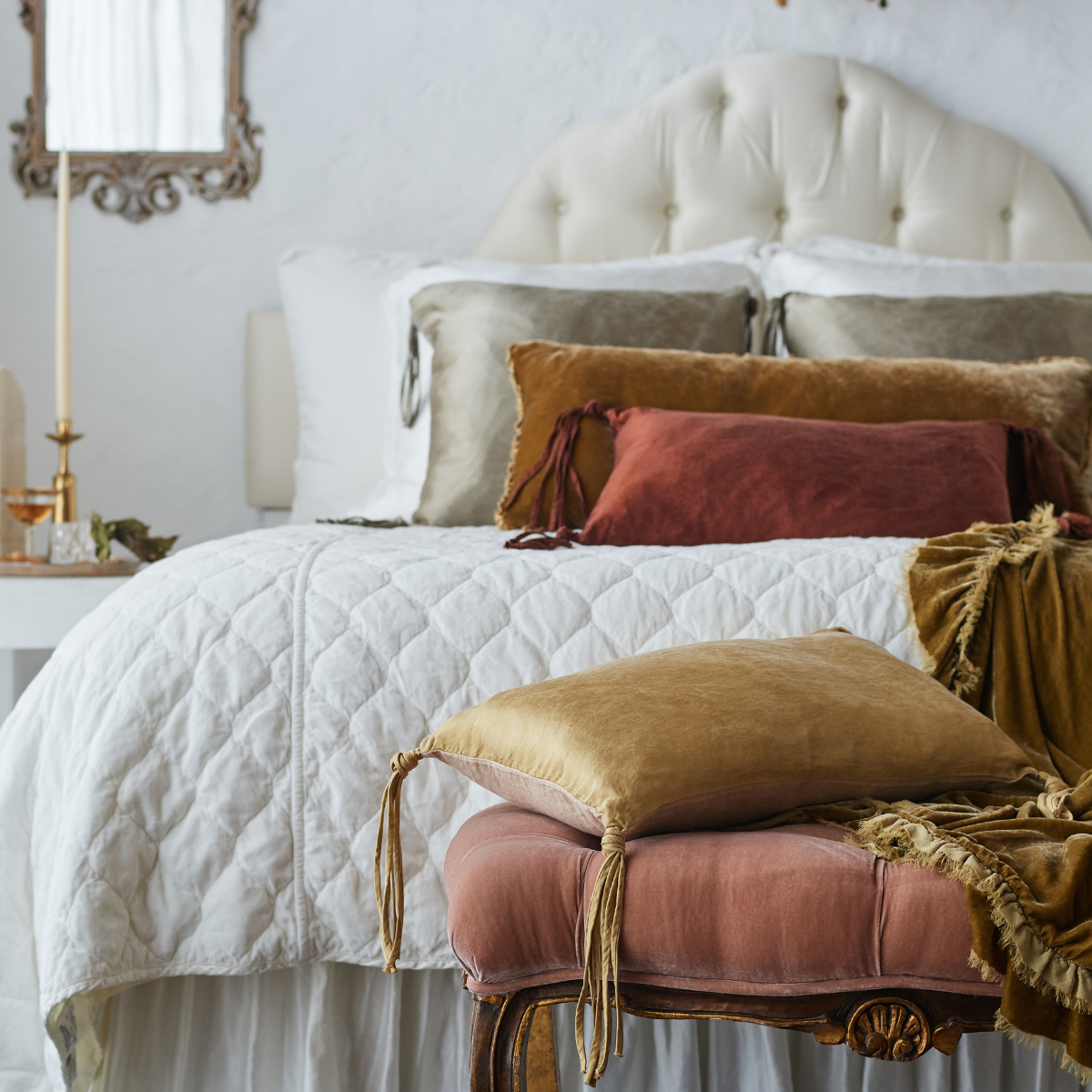 [allvariants]: Harlow coverlet in winter white on a neatly made bed against a grand tufted headboard. Warm brown and gold toned throw pillows and blankets in silk charmeuse and silk velvet provide contrast to the quilted cotton velvet - end of bed view. 