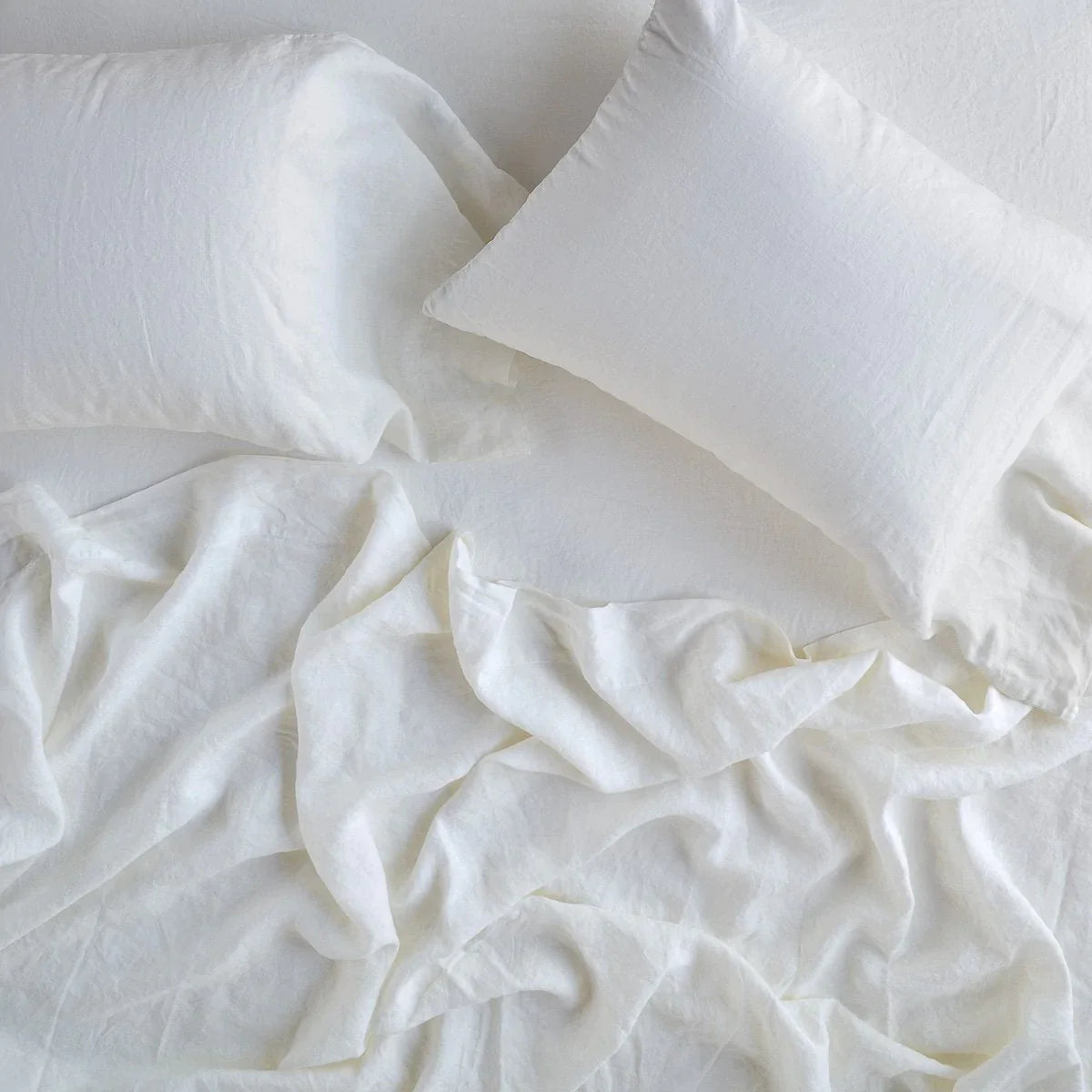 : Linen flat sheet in winter white, rumpled over a matching fitted sheet and sleeping pillows - overhead view.