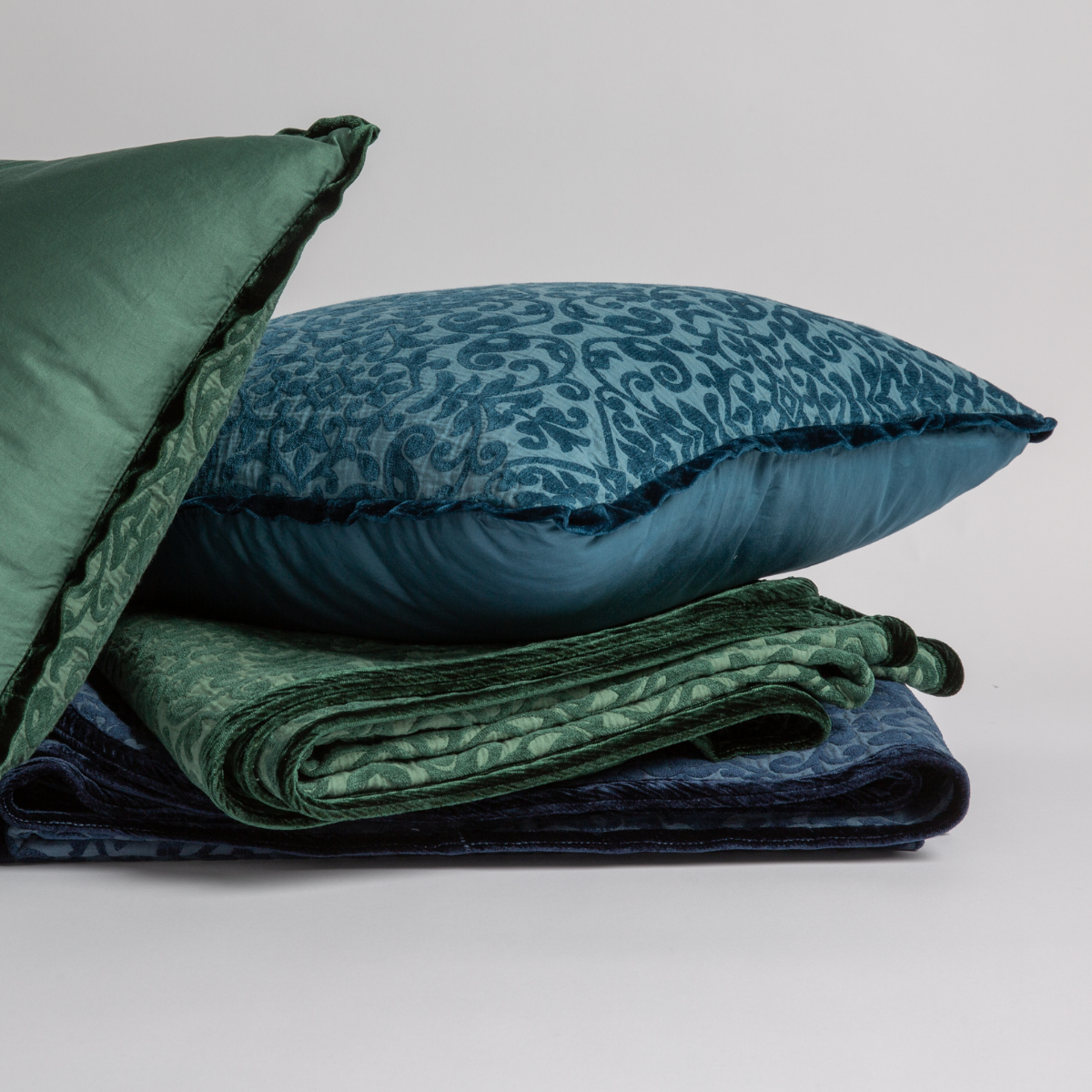 : Pillows in Jade and Cenote stacked on folded Vienna Throw Blankets in Jade and Midnight on a white background. 